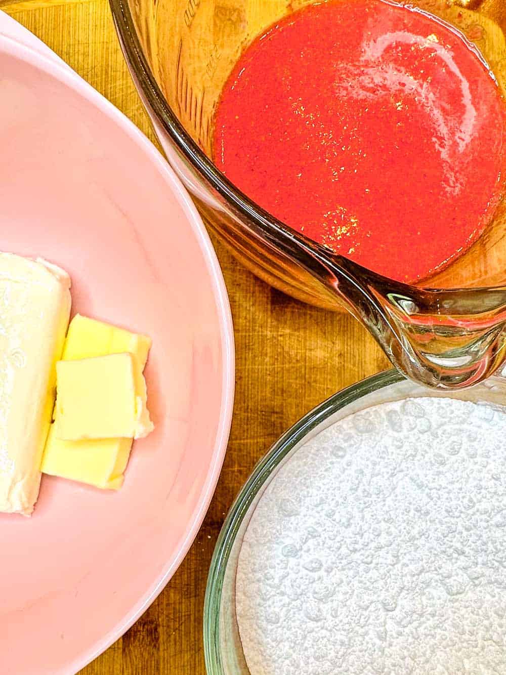 A pink bowl containing butter and cream cheese, a glass measuring cup filled with thick red liquid, and a glass bowl with powdered sugar are placed on a wooden surface, ready for baking preparation.