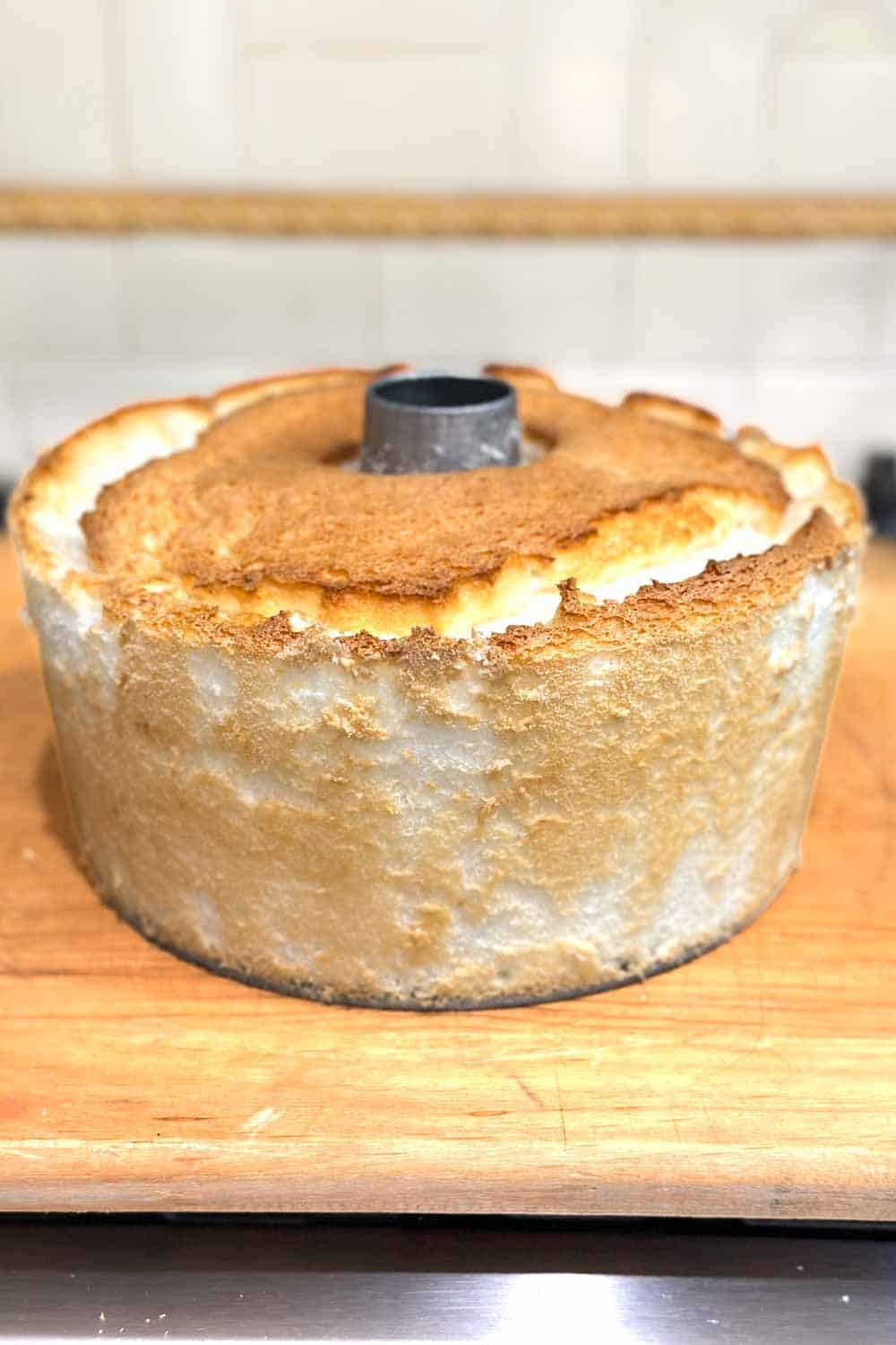 A freshly baked, golden-brown angel food cake is shown sitting on a wooden surface. The cake is in a traditional tube pan and has a fluffy, airy texture with a slightly cracked top. The sides of the cake are smooth and lightly browned from baking.