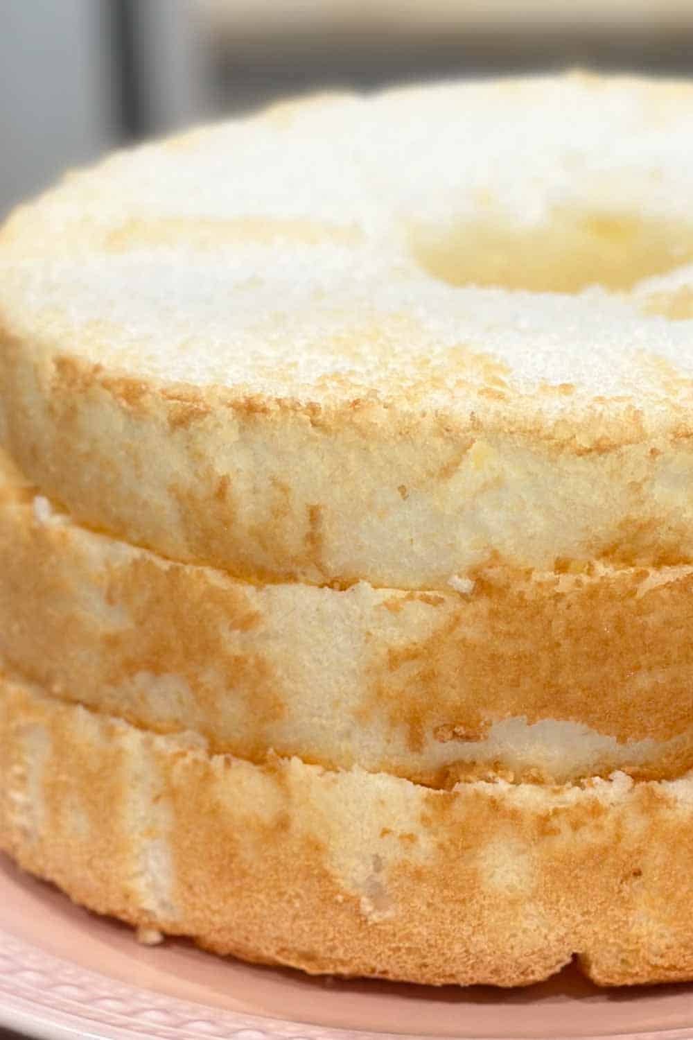 A close-up shot of a freshly baked angel food cake, showcasing its light and airy texture with a golden-brown, slightly crisp exterior. The cake is circular with a hole in the center, resting on a pink plate.