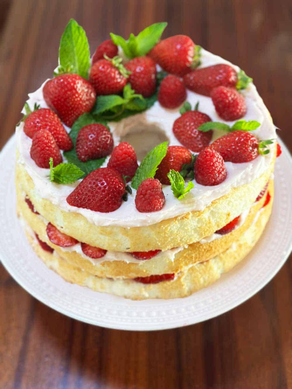 A layered sponge cake with a white icing top, garnished with fresh strawberries and mint leaves, is displayed on a white plate. The cake has visible layers of strawberries and cream in between. The background is a wooden surface.