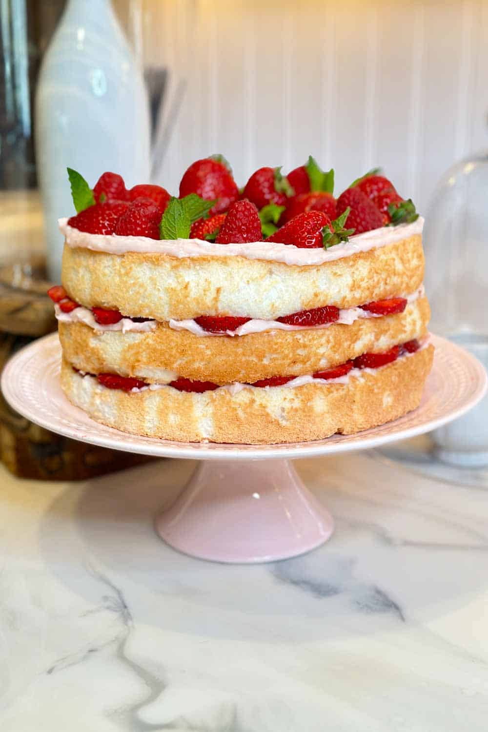 A layered sponge cake is topped with whole strawberries and garnished with mint leaves. The cake, set on a pink stand, features strawberry filling and frosting between each layer. The background shows a white counter and some blurred kitchen objects.