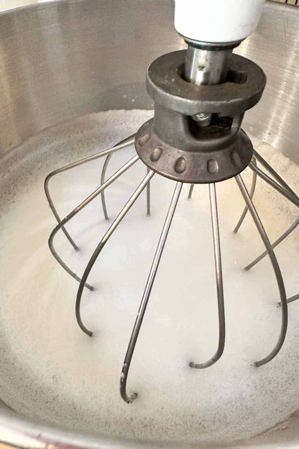 Close-up of a wire whisk attachment on a stand mixer, whipping egg whites in a stainless steel mixing bowl. The mixture is frothy, indicating the initial stages of beating the egg whites.