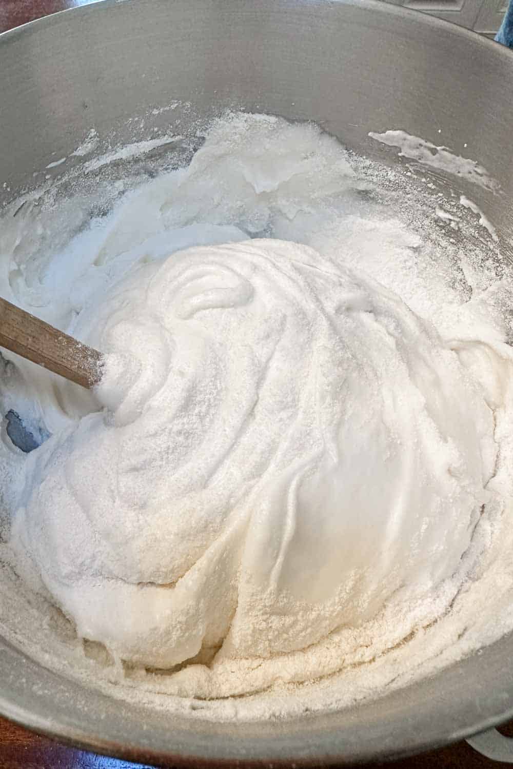 A large stainless steel mixing bowl filled with thick, white meringue. A wooden mixing spoon is partially submerged in the fluffy mixture. The meringue has soft peaks and is slightly glossy with a light dusting of powdered sugar visible on top.