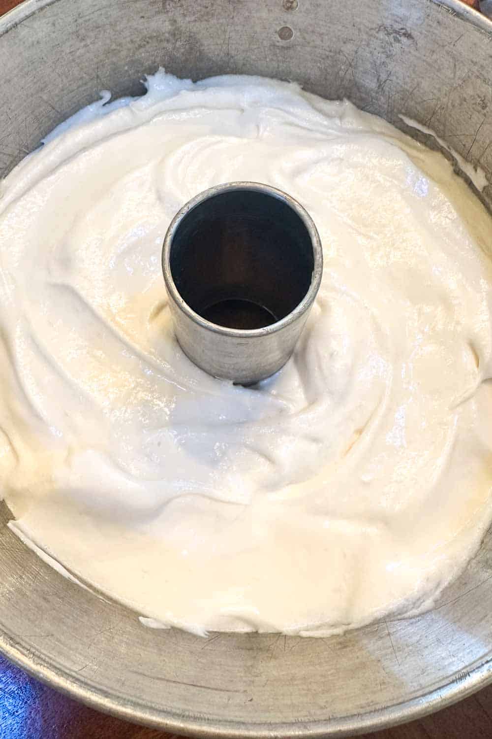 An unbaked angel food cake batter in a round tube pan. The batter is smooth and evenly spread, ready to go into the oven. The metal tube pan has a center hole for even baking.