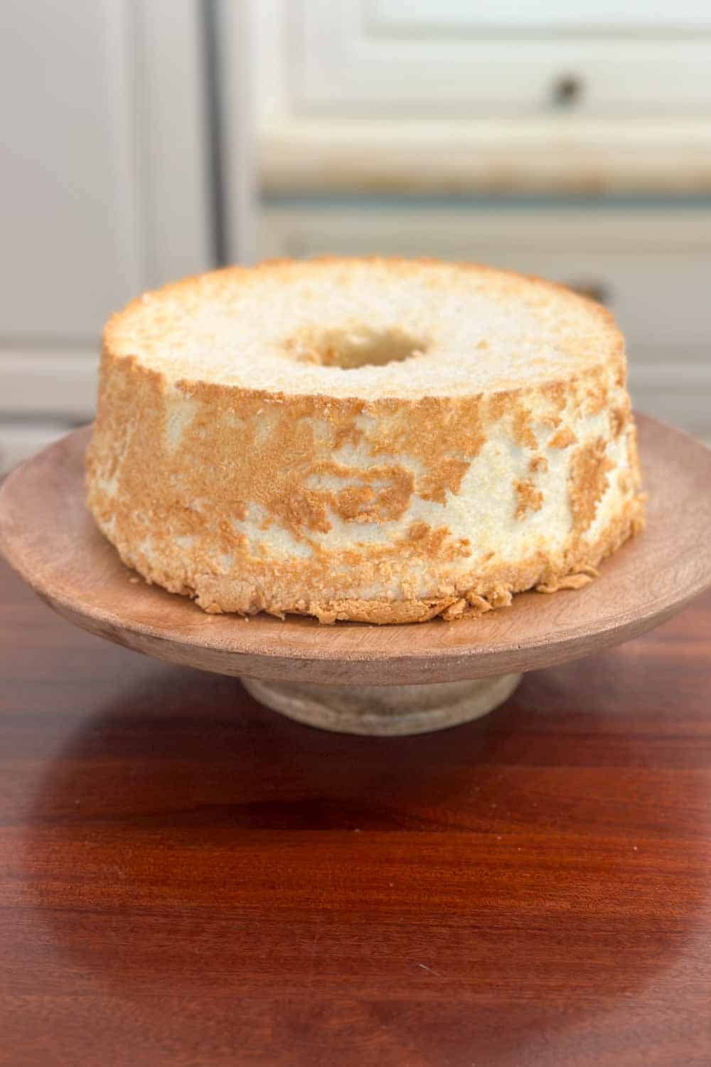 An angel food cake, golden brown on the outside, sits on a wooden cake stand. The cake is light and airy with a slightly textured exterior, and a hole in the center, characteristic of angel food cakes. The background is a wooden surface with a muted, light-colored wall.