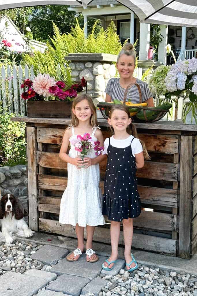 two girls and a grandmother standing at a wood pallet bar selling flowers