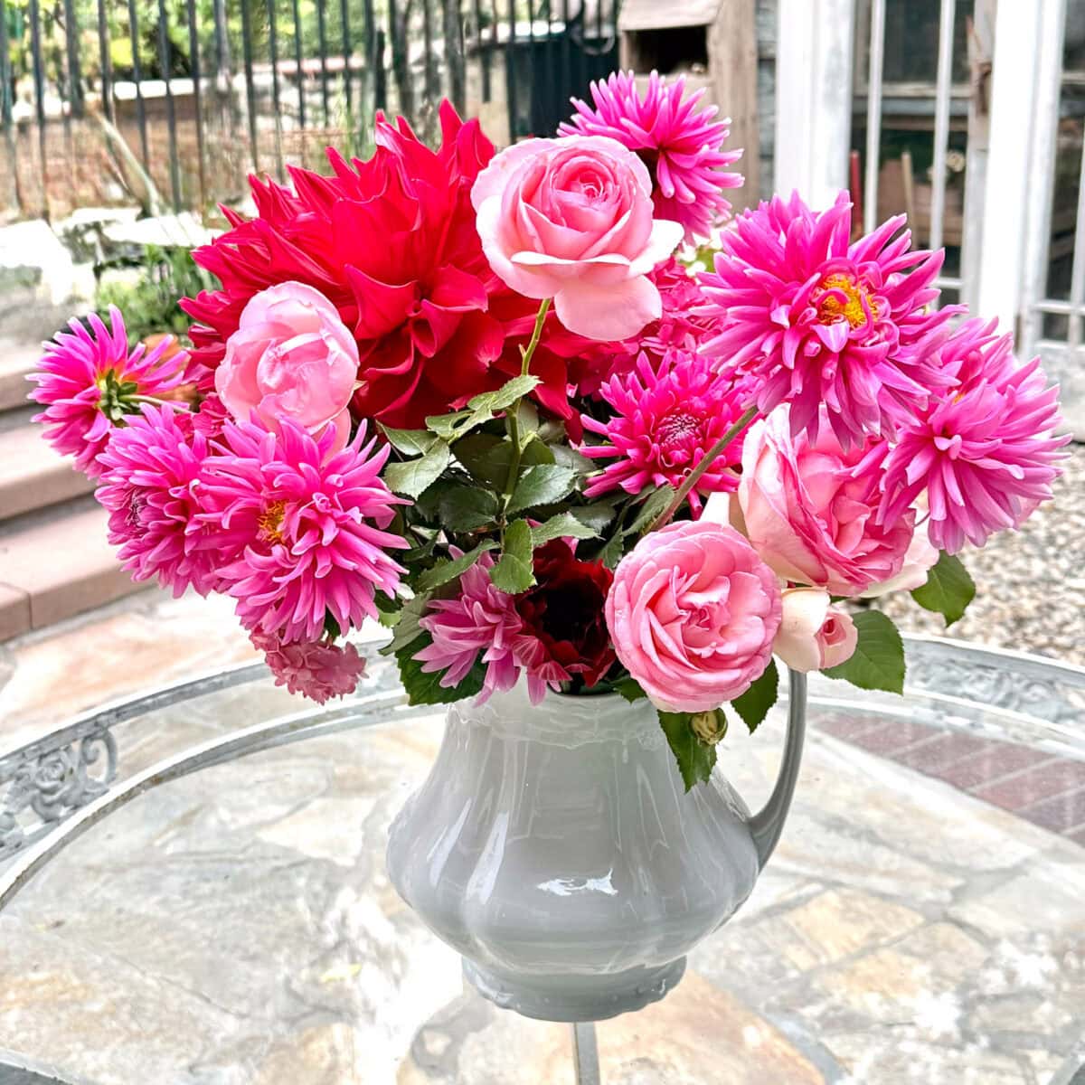 A white ceramic pitcher holding a bouquet of pink dahlias and roses sits on a glass table outdoors, showcasing elegant dahlia flower arrangements with stone steps and a garden in the background.