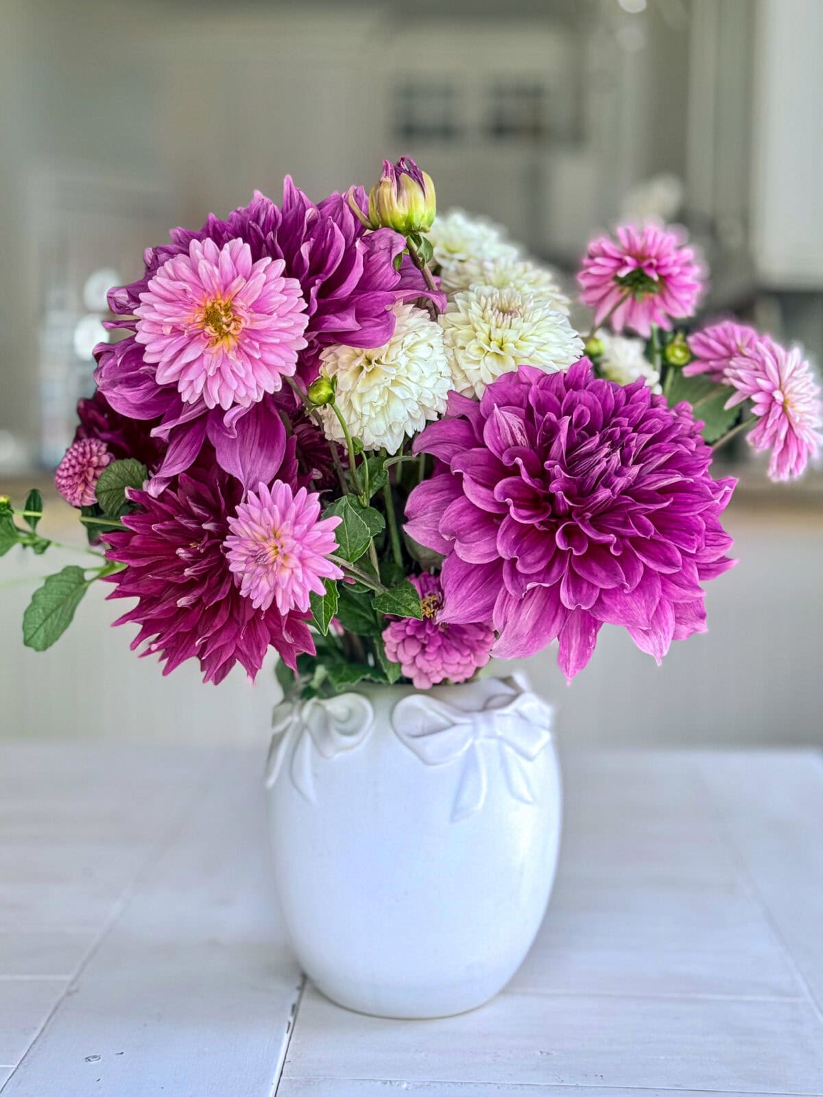 A white vase with a bow design holds a bouquet of purple, pink, and white dahlias on a white table with a blurred kitchen background, showcasing elegant dahlia flower arrangements.