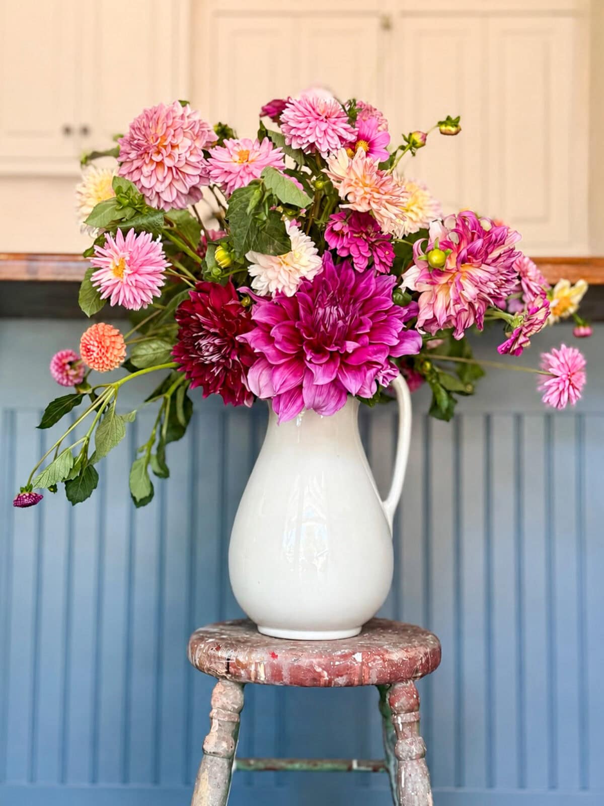 A white ceramic pitcher filled with pink and purple dahlia flower arrangements sits on a weathered wooden stool in front of a blue paneled wall and white cabinets.
