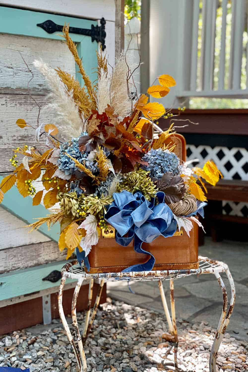 A decorative fall arrangement in a vintage suitcase sits on an ornate metal stand. The arrangement features autumn leaves, dried pampas grass, blue hydrangeas, berries, and a large blue ribbon. The suitcase is against a rustic wooden backdrop with a turquoise door.