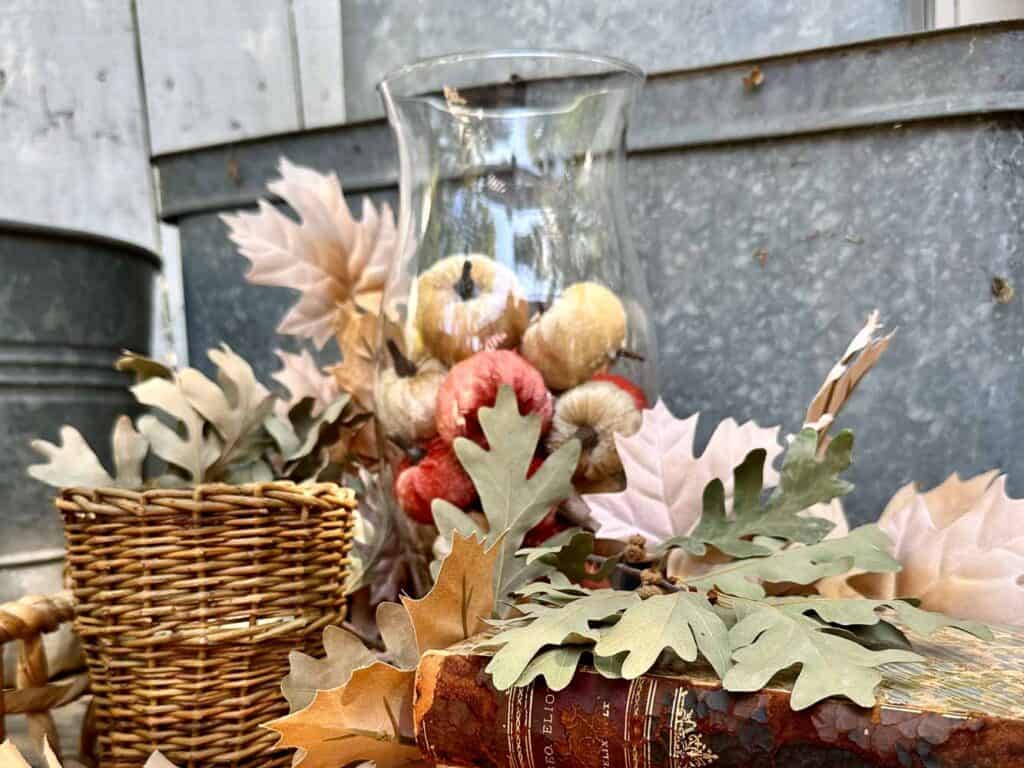 Fall vignette with a hurricane glass vase full of small velvet pumpkins, a wicker candle and vintage books with fall leaves