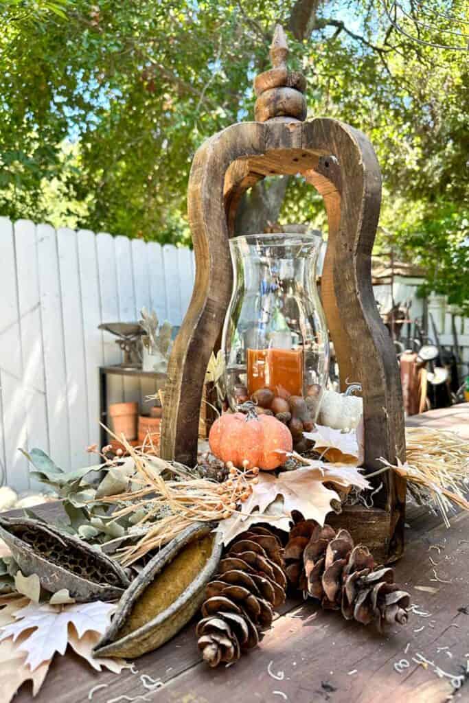 Wooden open lantern with a glass hurricane lamp filled with a candle and acorns. A pumpkin and fall leaves finish the look.