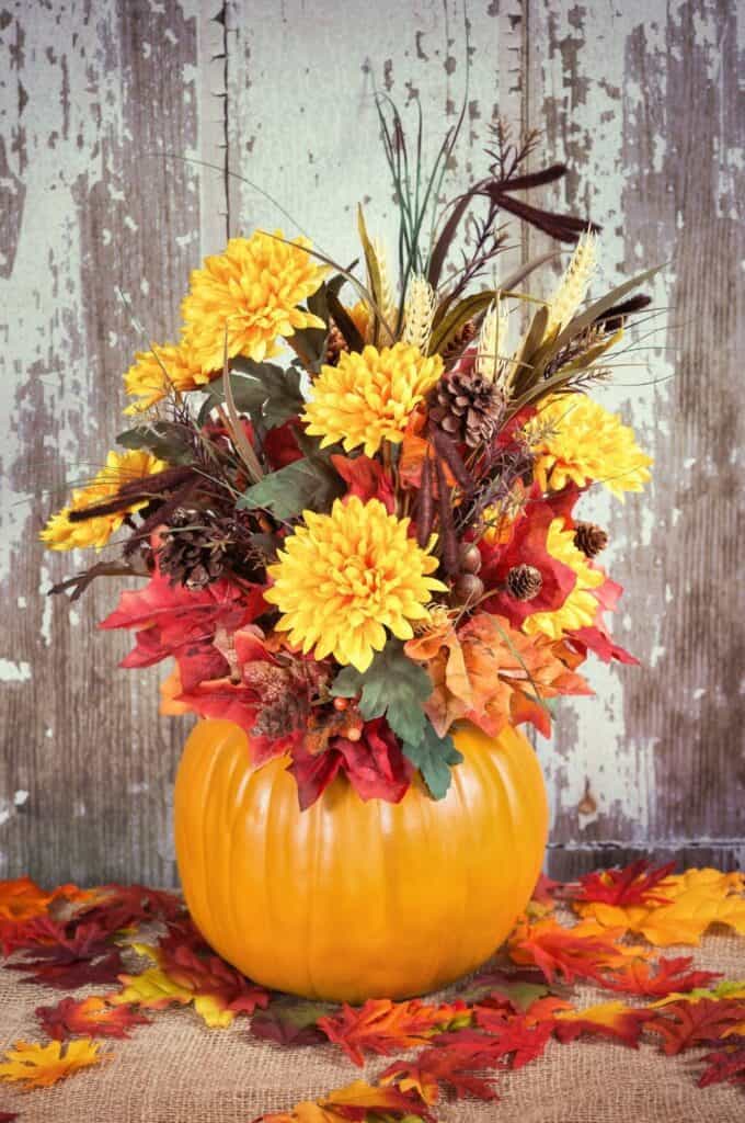 A vibrant autumn arrangement featuring yellow chrysanthemums, red and orange leaves, pinecones, and wheat stalks, displayed in a hollow pumpkin vase. Perfect for front porch fall decorating ideas on a budget, it's set on a rustic wooden background with scattered fall leaves on the burlap-covered table.