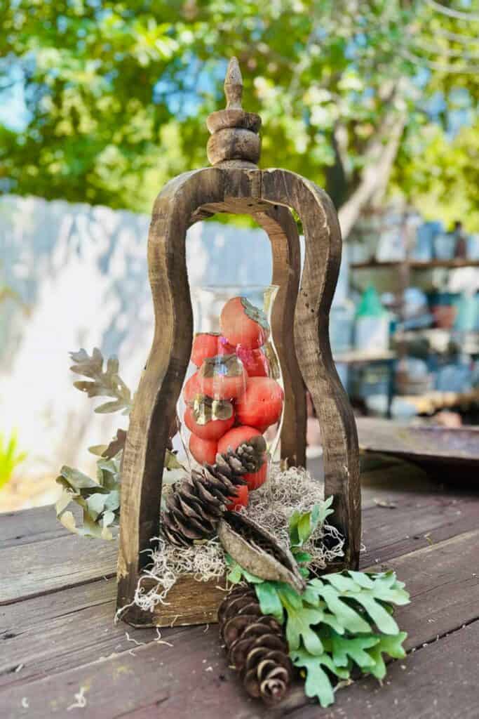 Wood lantern with a glass hurricane filled with persimmons and pine cones for a fall decoration. 