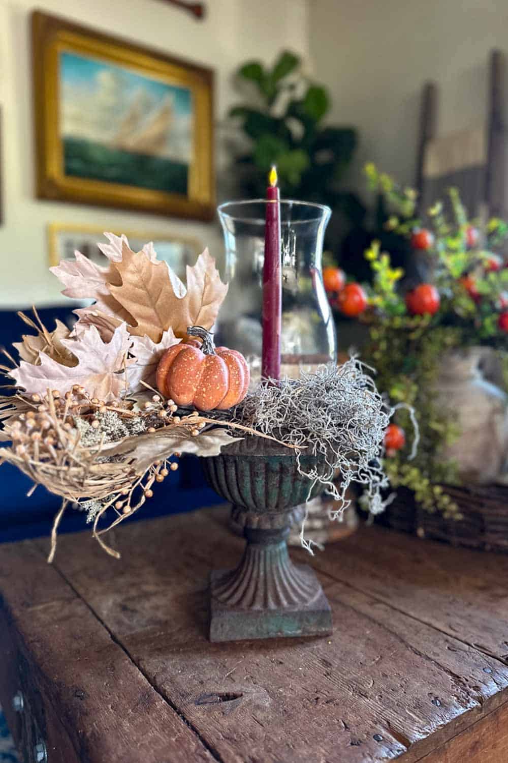 Glass hurricane vase centerpiece sitting in a vintage urn holding a single taper candle and some faux fall leaves with a pumpkin. 