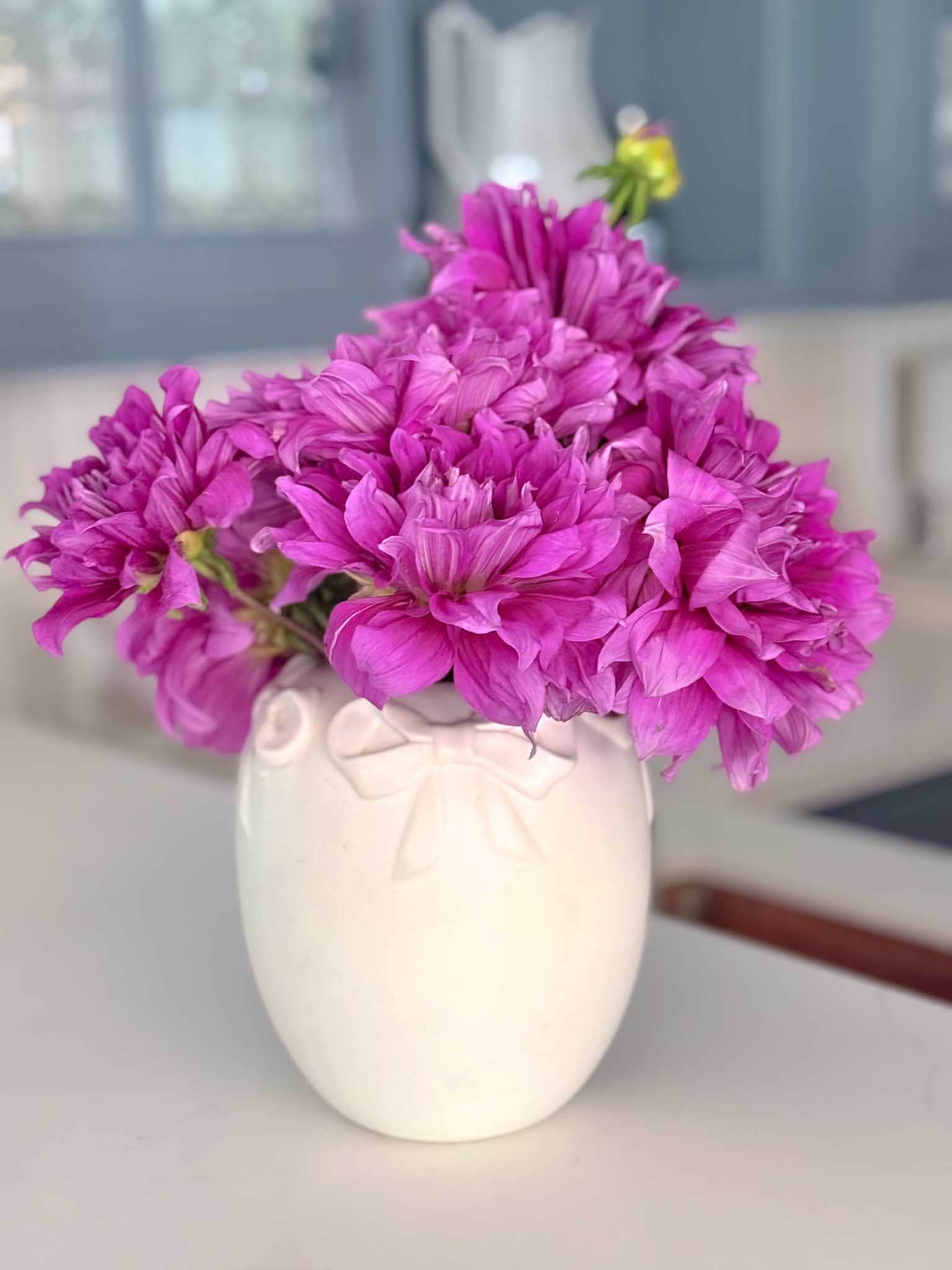 A white vase with a bow design holds a bouquet of vibrant purple dahlia flower arrangements, set on a light-colored surface with a blurred kitchen background.