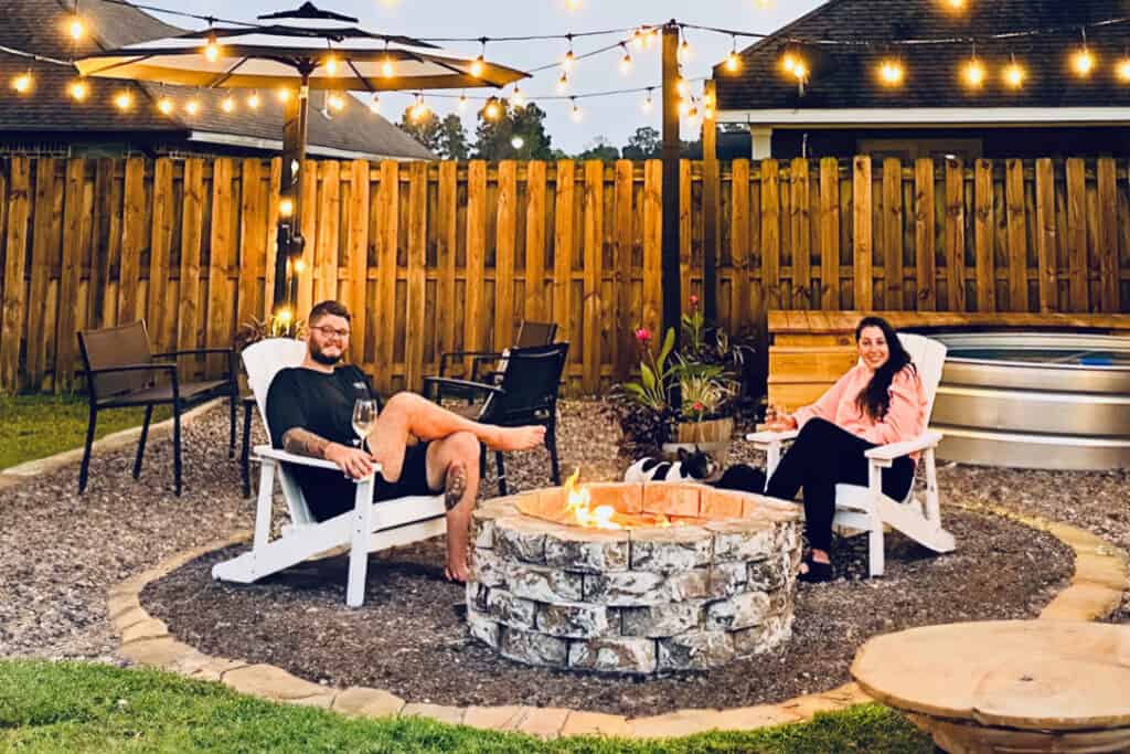 Two people sitting in white Adirondack chairs around a circular stone fire pit in a backyard with a wooden fence. String lights are hanging overhead, and a hurricane vase for fall decorates the scene, creating a cozy atmosphere. Both are smiling and holding drinks, with more seating and an umbrella in the background.