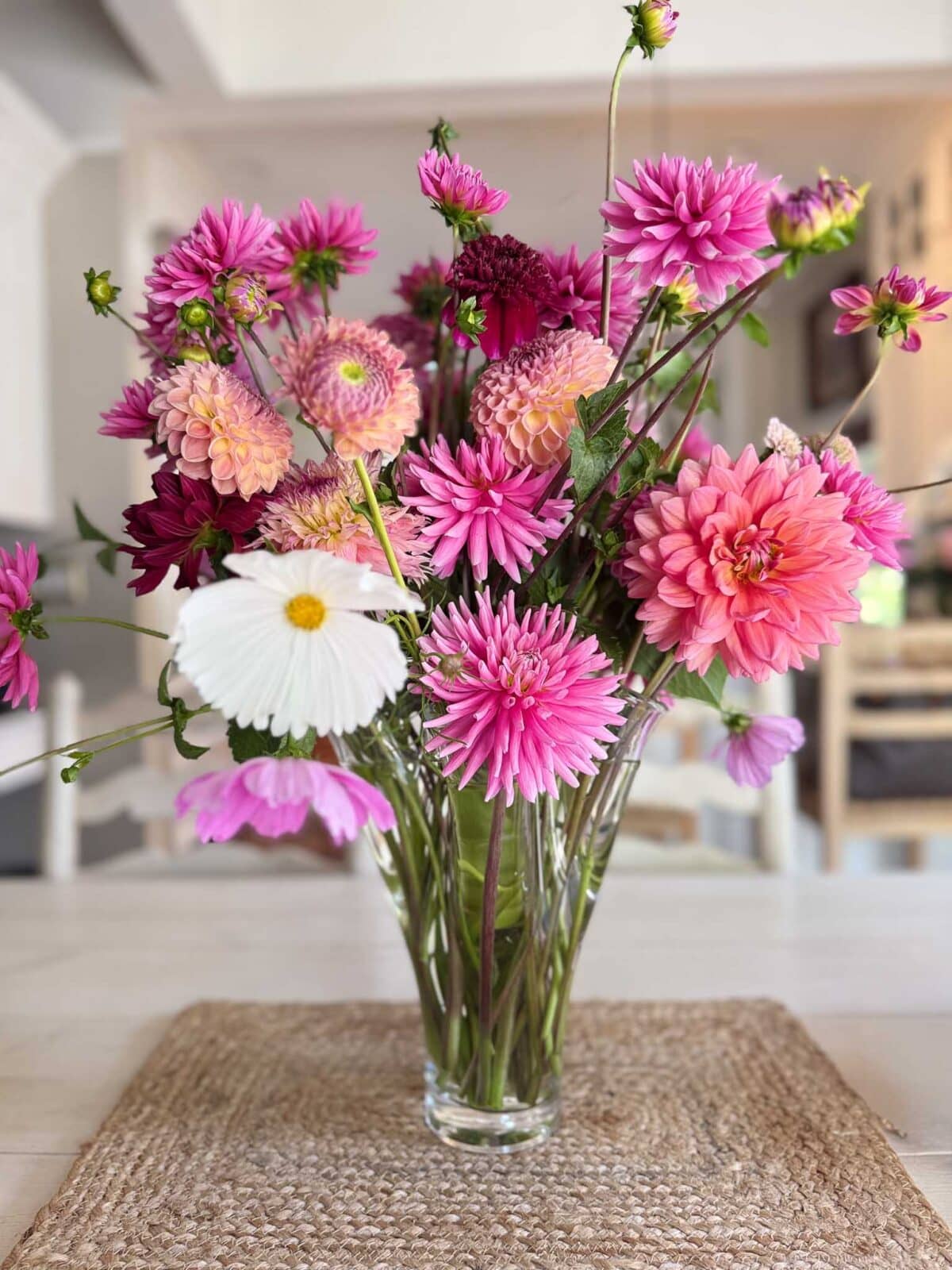 A clear glass vase holds a bouquet of pink, purple, and white dahlia flower arrangements with green stems, placed on a woven mat atop a light-colored table in a bright indoor setting.