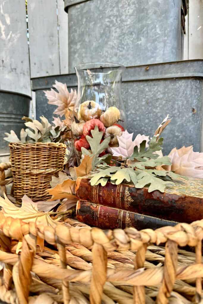 Fall vignette with a hurricane glass vase full of small velvet pumpkins, a wicker candle and vintage books with fall leaves