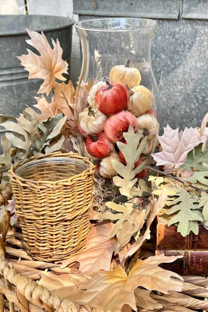 Fall vignette with a hurricane glass vase full of small velvet pumpkins, a wicker candle and vintage books with fall leaves