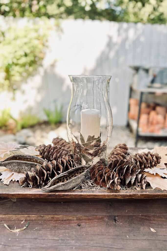 Simple fall arrangement with pine cones and a candle wrapped with leaves and small pine cones inside of a hurricane glass vase sitting in a dough bowl.