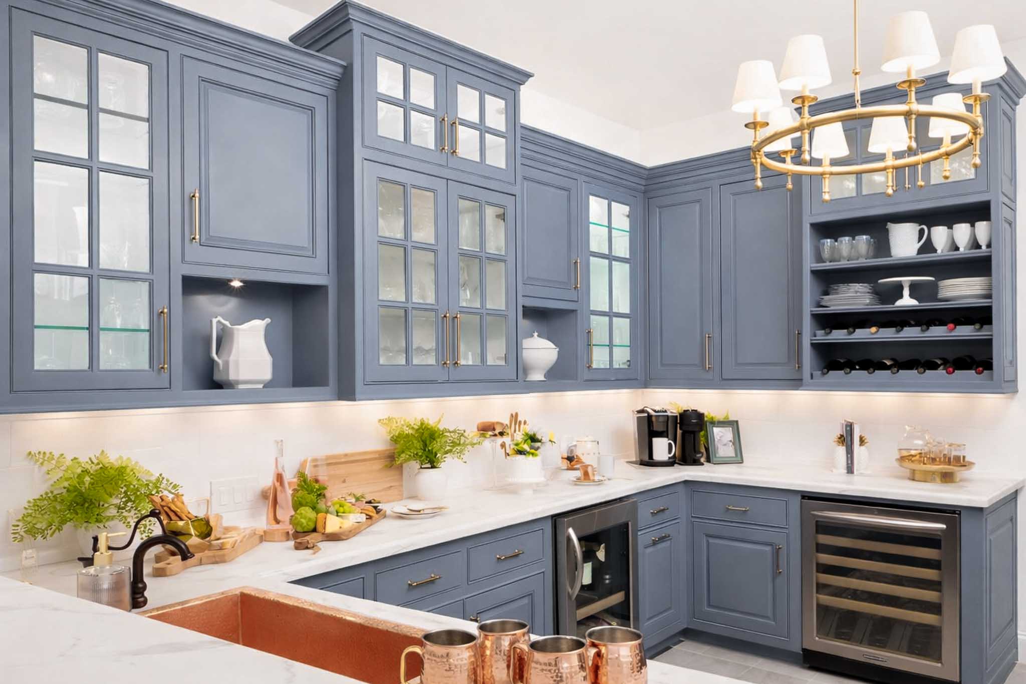 Modern kitchen featuring the best blue gray for kitchen cabinets, glass-front doors, white countertops, built-in wine rack and fridge, copper sink, gold light fixture, plants, and stylish kitchenware on counters and shelves.