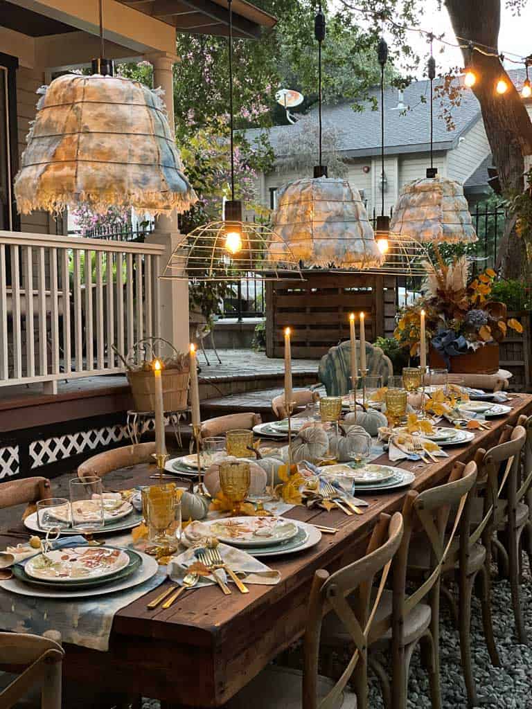 Candles on a fall table outside. The table is set with blue and gold linens, with hanging light fixtures over the table. Pretty fall dishes with pumpkins adorn the table.