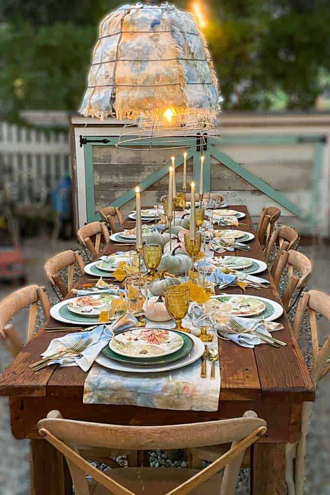 A rustic outdoor dining table, embodying fall tablescape ideas, is set for a meal with wooden chairs. The table features tall candles, plates with patterned napkins, yellow and clear goblets, and decorative pumpkins. A unique fabric-covered hanging light fixture illuminates the scene.