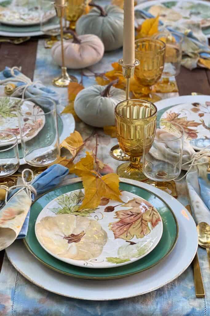 Place setting at a fall harvest table. A cream-colored charger plate with a green dinner plate and a pumkin design on the salad plate. Amber glasses and gold silverware accent the table nicely. 