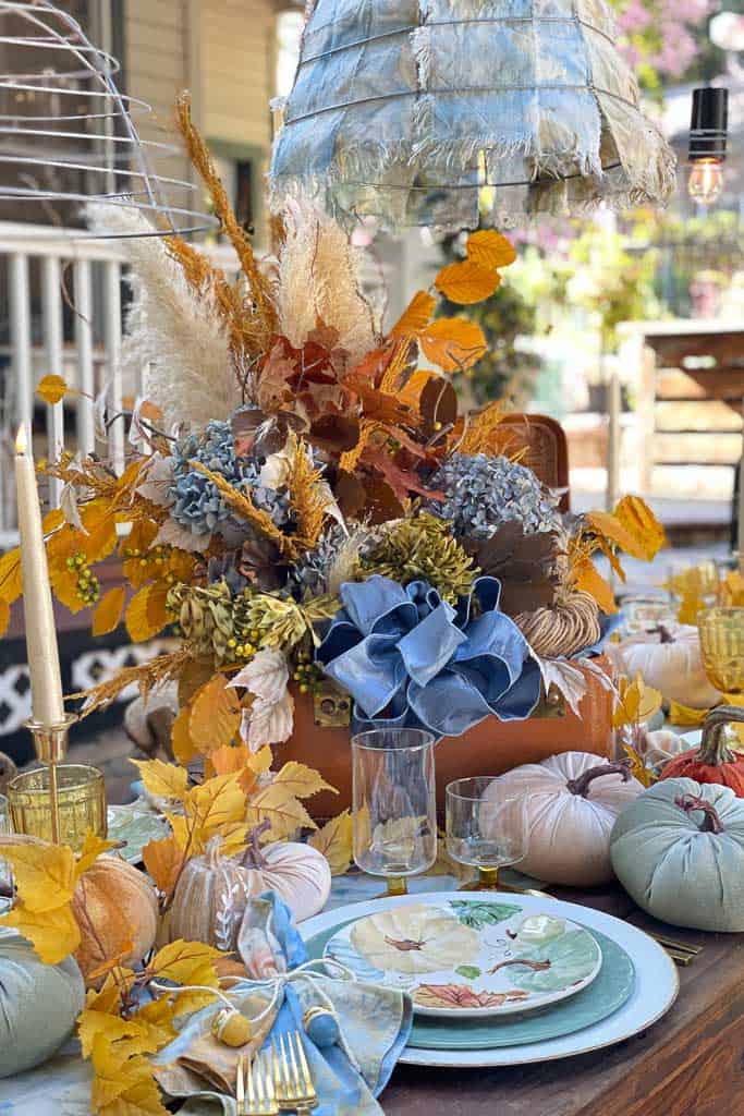Outdoor fall tablescape with a large centerpiece that is all blue and gold. This centerpiece is made of dried flowers and sitting in a vintage train case. The table is also adorned with pumpkins and pretty fall plates.