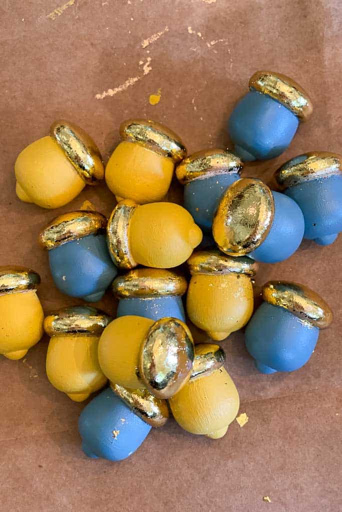 A collection of small acorn decorations lies on a brown surface. The acorns are painted in alternating shades of yellow and blue, each topped with a shiny, metallic gold cap.