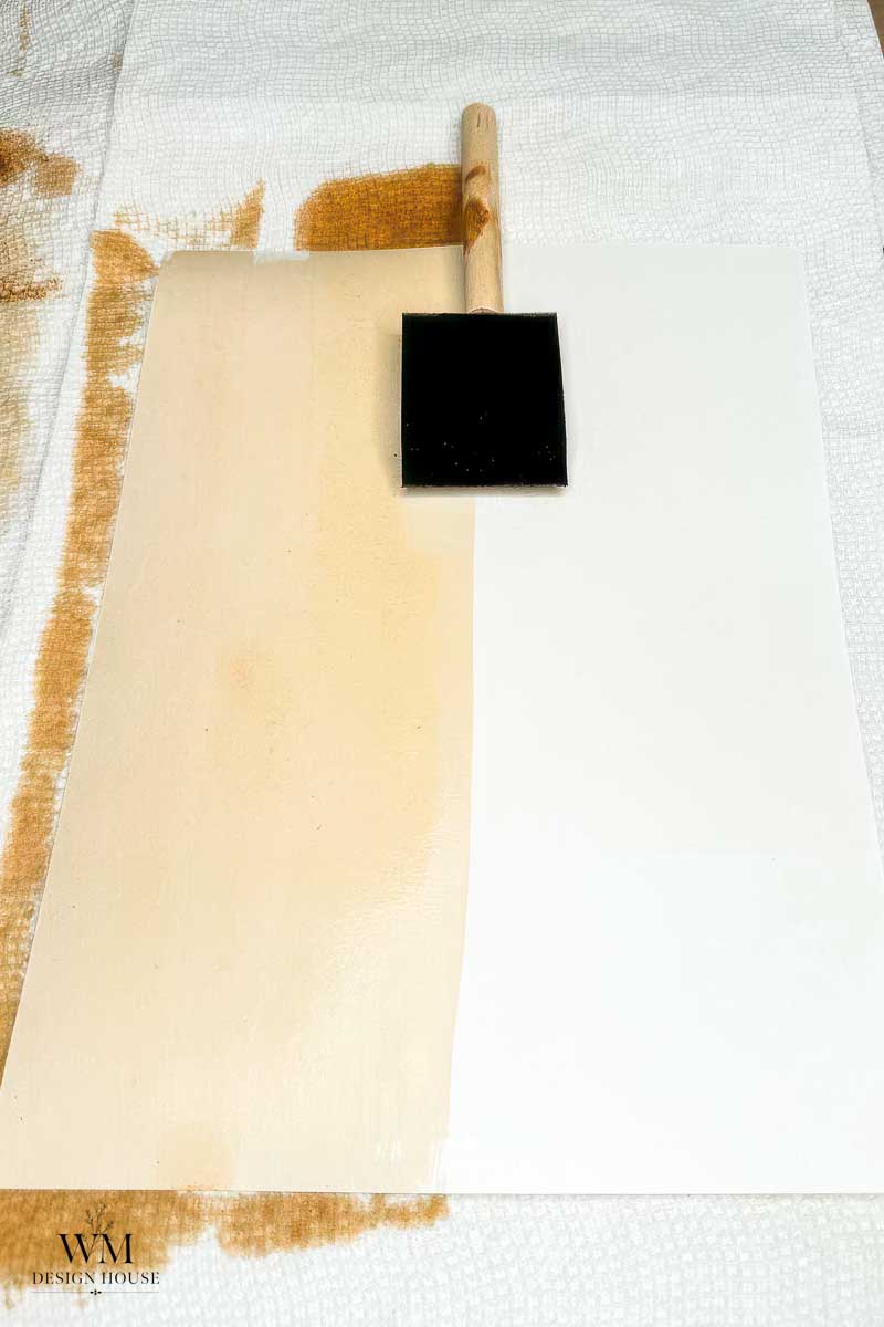 A foam brush rests on a sheet of paper, half stained a light tan, half white—an essential step in learning how to make coffee paper. The brush lies across the division of colors, while paper towels protect the surface underneath.