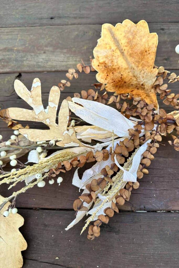 A close-up of a rustic autumn-themed decorative arrangement on a wooden surface. It features various dried leaves in shades of beige and brown, small clusters of dried seeds, and white berries, creating a warm, seasonal aesthetic.