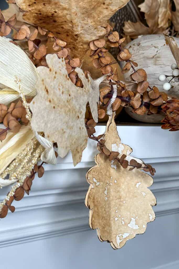 A close-up of an autumn-themed decoration featuring dried leaves, brown eucalyptus branches, and ornamental accents in neutral tones. The arrangement is set on a white surface, highlighting the textures and natural beauty of the dried foliage.