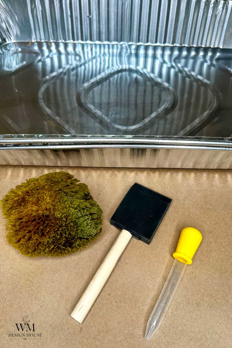 A metal tray, a sea sponge, a foam paintbrush with a wooden handle, and a plastic dropper with a yellow bulb are arranged on a brown surface—tools often gathered for to make coffee stained paper.
