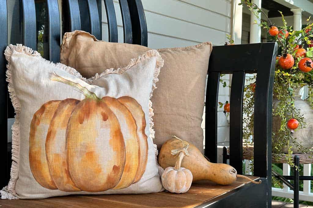 Painted pumpkin pillow sitting on a black bench on the front porch with gourds and one other solid-colored pad.