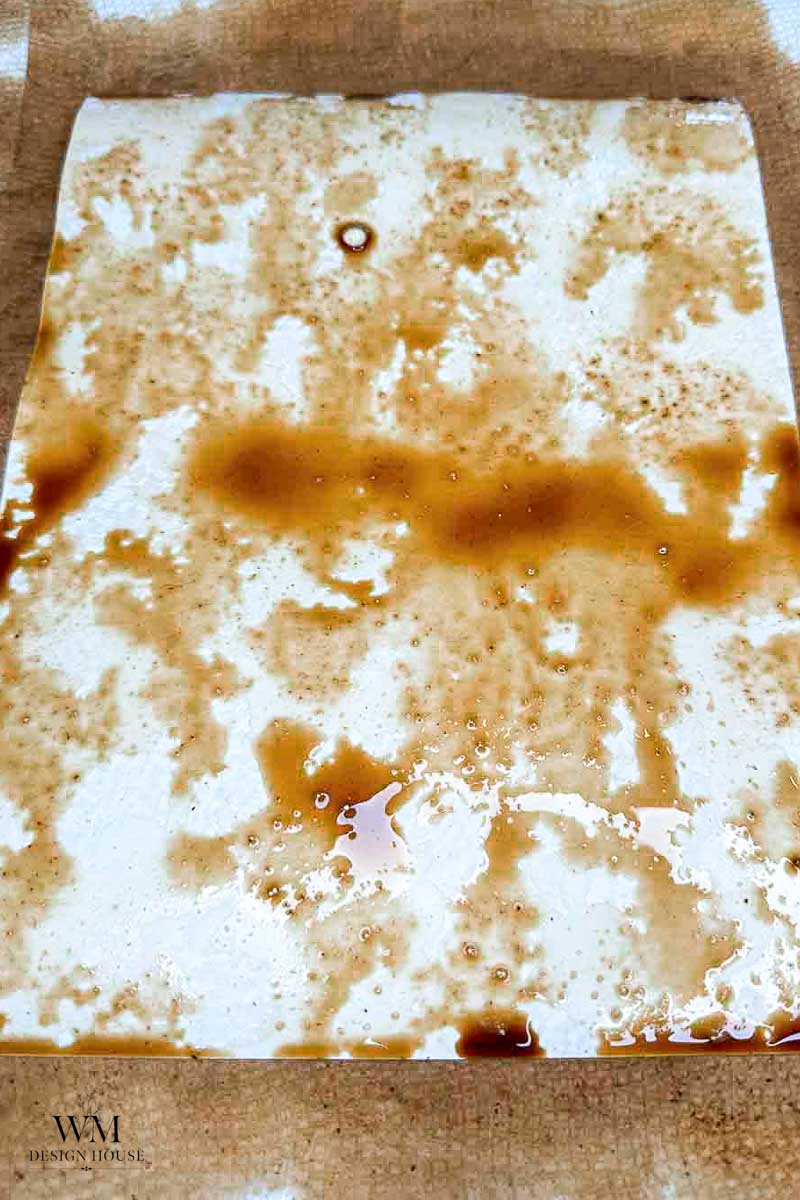 A sheet of white paper with scattered brown stains, possibly from coffee or tea, lies on a textured beige surface. The marks are irregular and spread across the page, showing the result of how to make coffee paper by staining it for a vintage effect.