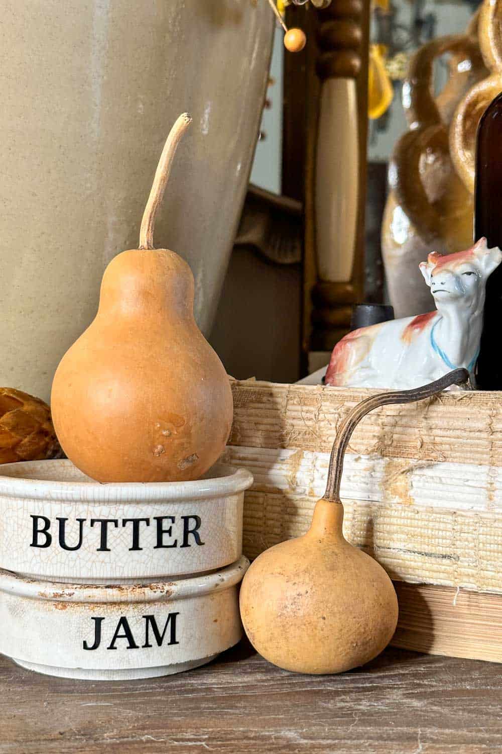 Two small gourds rest atop stacked ceramic containers labeled "BUTTER" and "JAM." A decorative cow figurine and various objects in warm tones adorn the background, creating a rustic and cozy atmosphere.
