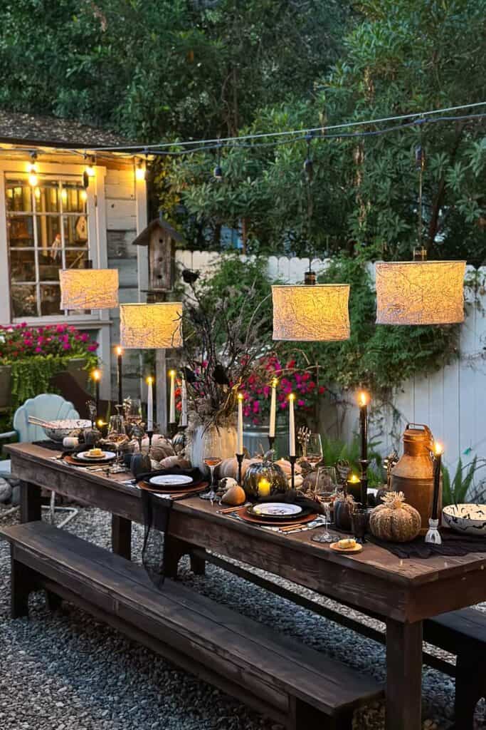 A beautifully decorated outdoor table with autumn-themed and Halloween decor, featuring pumpkins, candles, and rustic tableware. Four hanging lamps illuminate the setting, surrounded by greenery and a cozy, inviting atmosphere perfect for a festive Halloween gathering.