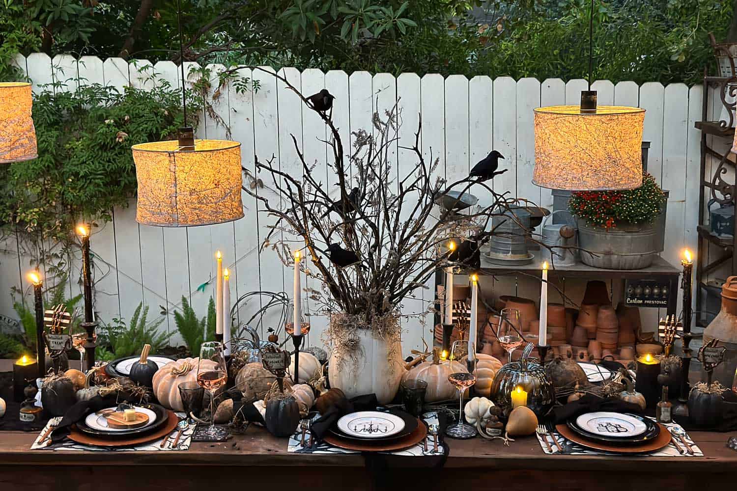 Halloween outdoor tablescape with a spooky tree and crows as the centerpiece. Shades of gray illuminate the table with lots of pumpkins and rusty elegance. Dishes are black and white, and the name cards are held with a skeleton hand. 