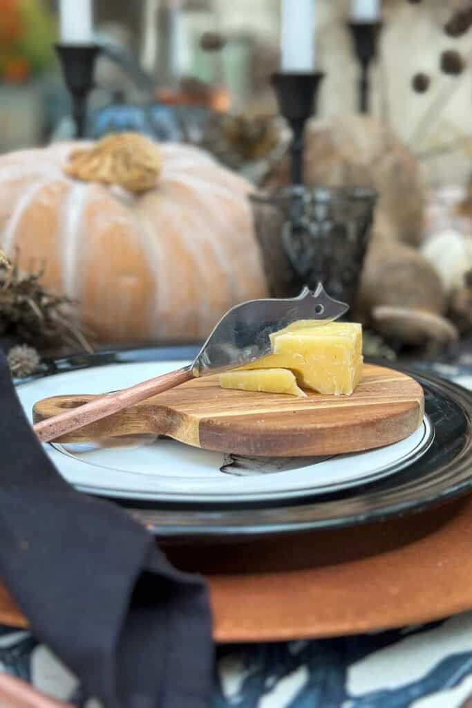 A decorative dining setup featuring a wooden cutting board with cheese and a cheese knife on top of stacked plates. A black napkin is placed beside the plate, and a pumpkin and candles are visible in the blurred background, evoking an autumn theme.