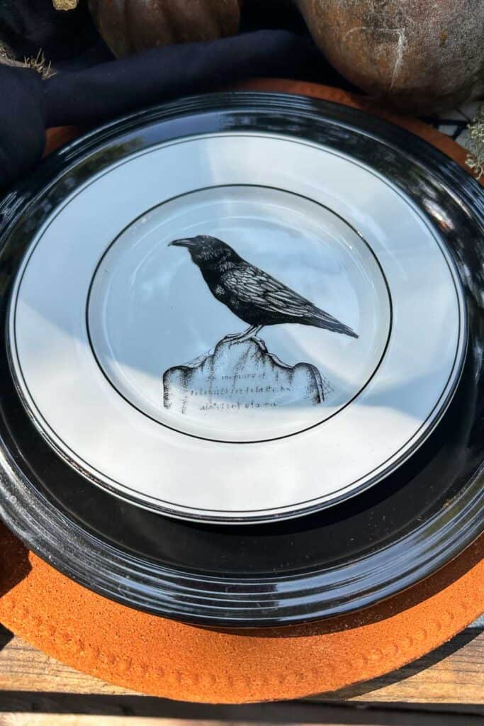 A white plate with a black rim features an illustration of a black crow perched on a rock. The plate is set on top of a larger black plate and an orange placemat. The background includes a rustic setting with a partially visible pumpkin.