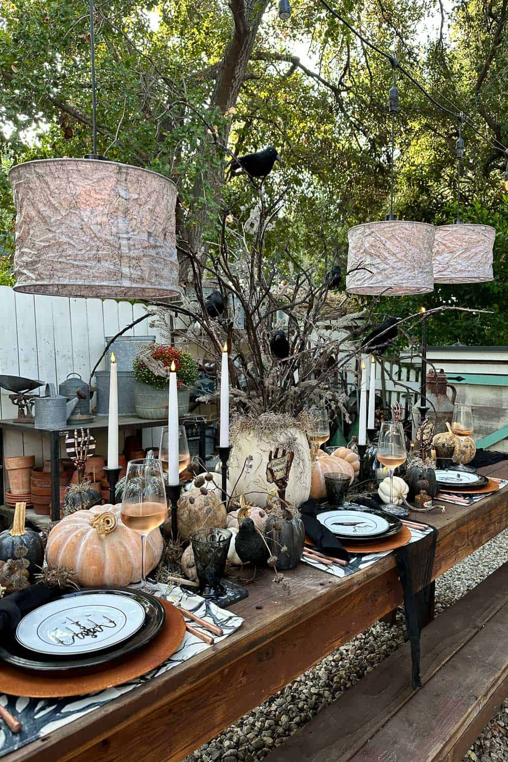 Halloween outdoor tablescape with a spooky tree and crows as the centerpiece. Shades of gray illuminate the table with lots of pumpkins and rusty elegance. Dishes are black and white, and the name cards are held with a skeleton hand. 