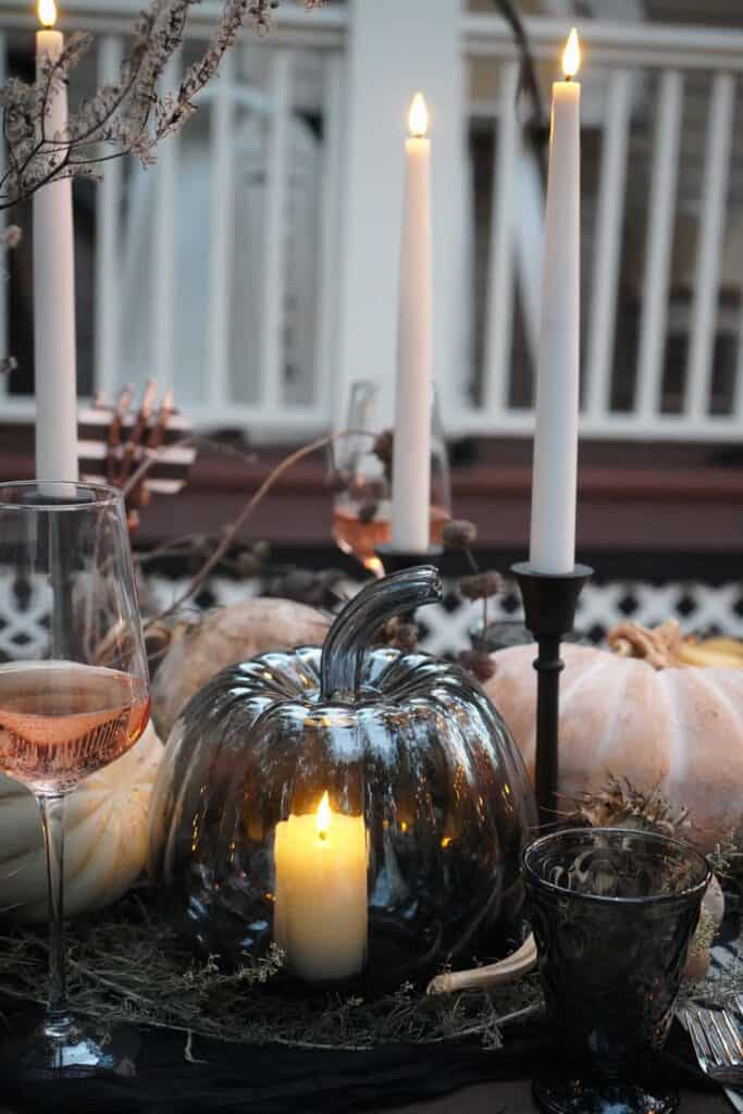 A table is elegantly decorated with a dark metallic pumpkin candle holder illuminating a candle, surrounded by white pumpkins, tall lit taper candles in black holders, moss, and wine glasses filled with rosé wine, creating a cozy, festive atmosphere.