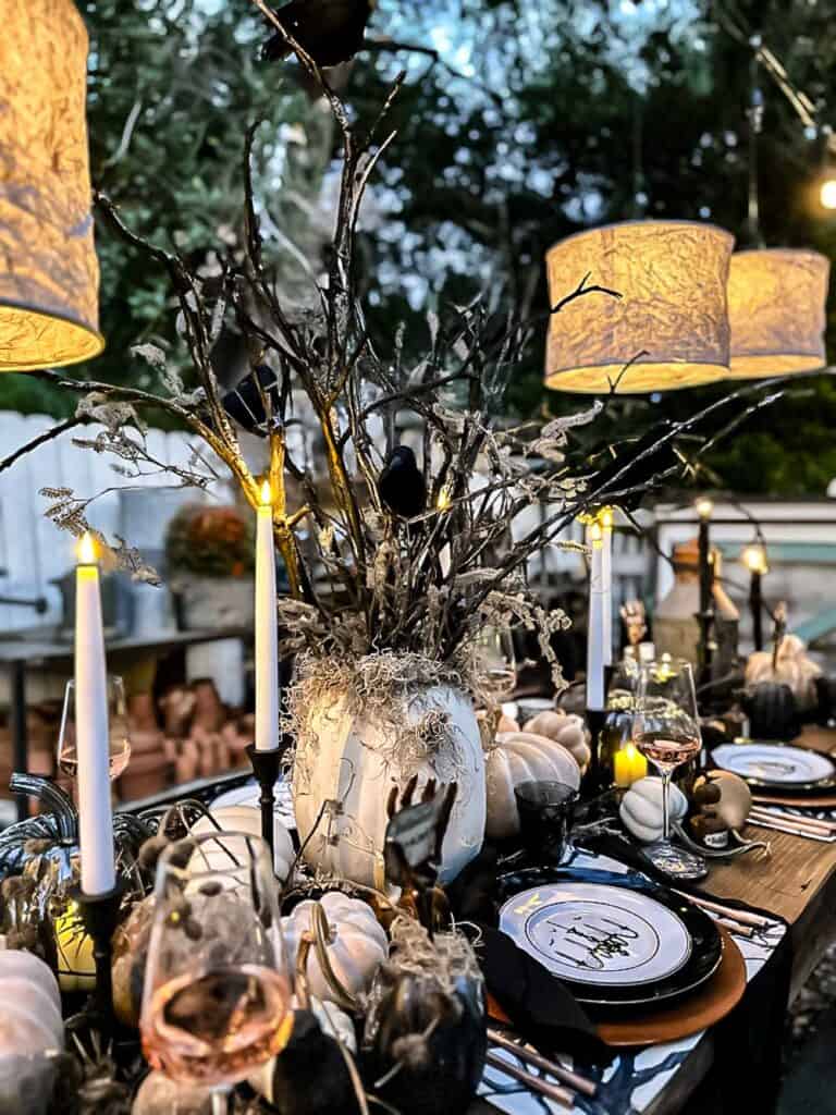 The Halloween table is adorned with a DIY touch: imaginative place card holders complement the centerpiece of branches and birds. White pumpkins, flickering candles, and elegant settings blend seamlessly into the décor, while warm hanging lamps cast a cozy yet spooky glow over the festive Halloween decorations.