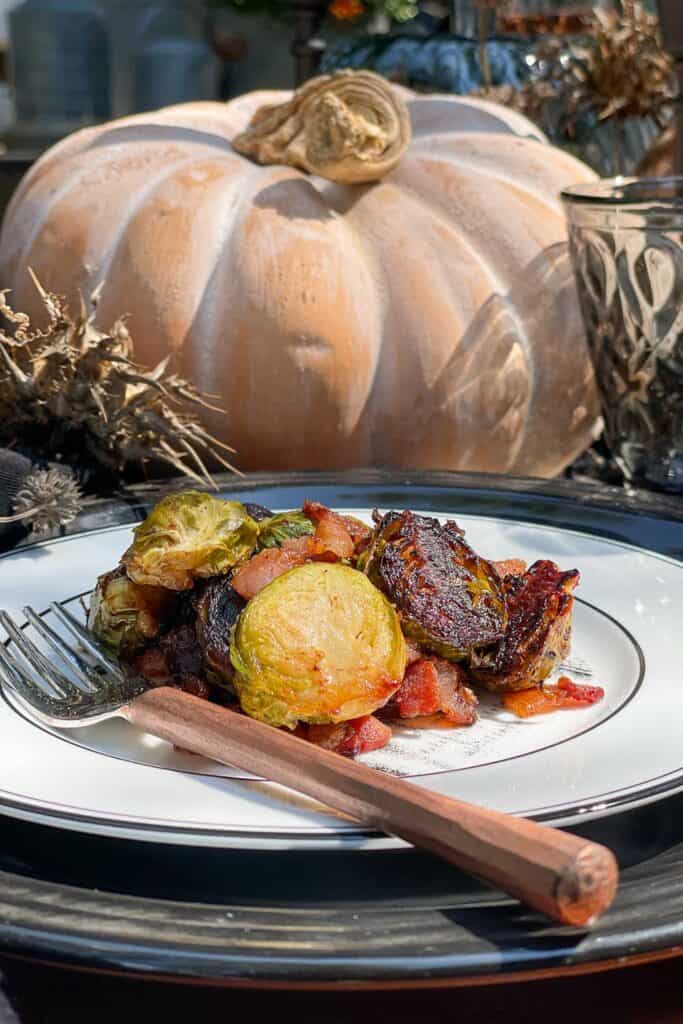 Roasted Brussels sprouts served on a plate with dates and bacon. The black and white plate is sitting on a table with fall pumpkins.