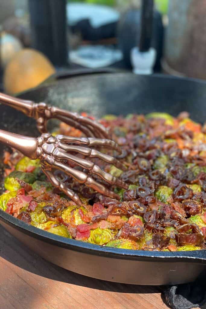 A cast iron pan with Brussels sprouts, bacon, and dates sitting on a table with black candles and other fall/ Halloween decor.