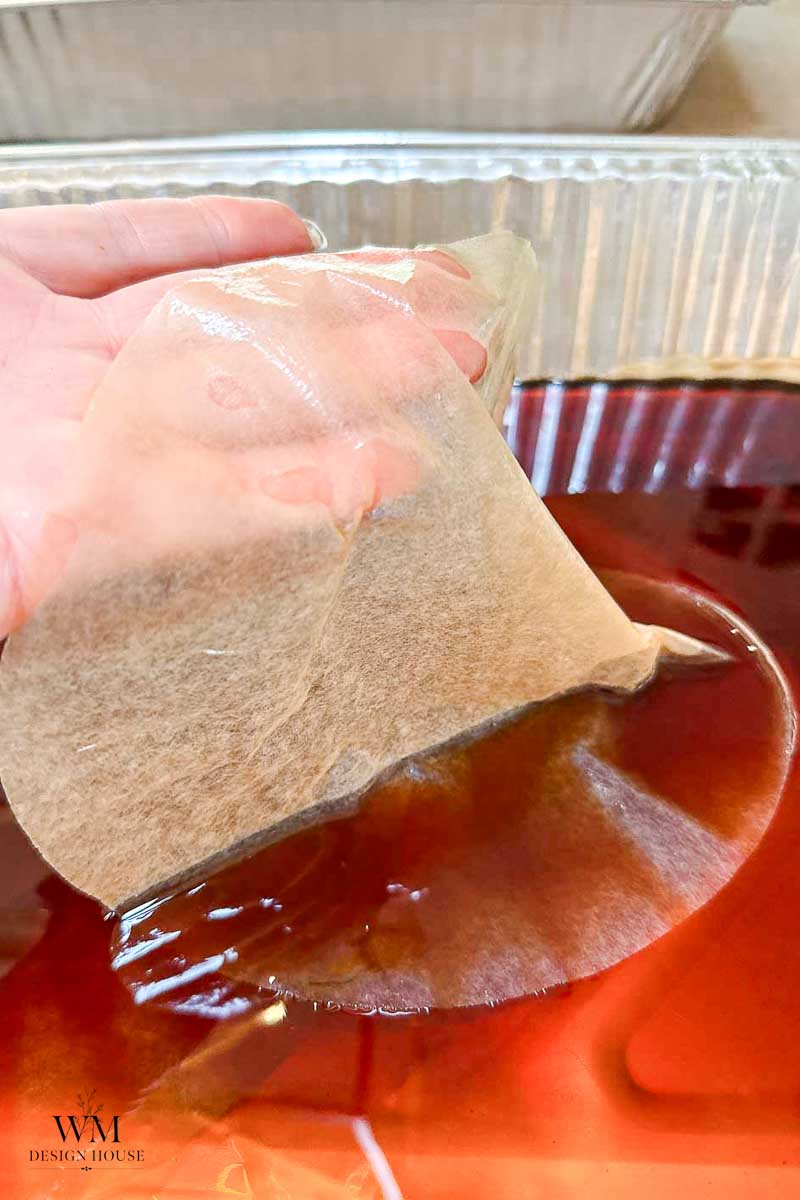 A hand lifts a translucent sheet of paper from a pan filled with dark reddish liquid, likely dye or tea, for a crafting or aging process—demonstrating how to make coffee paper with rich, vintage tones.