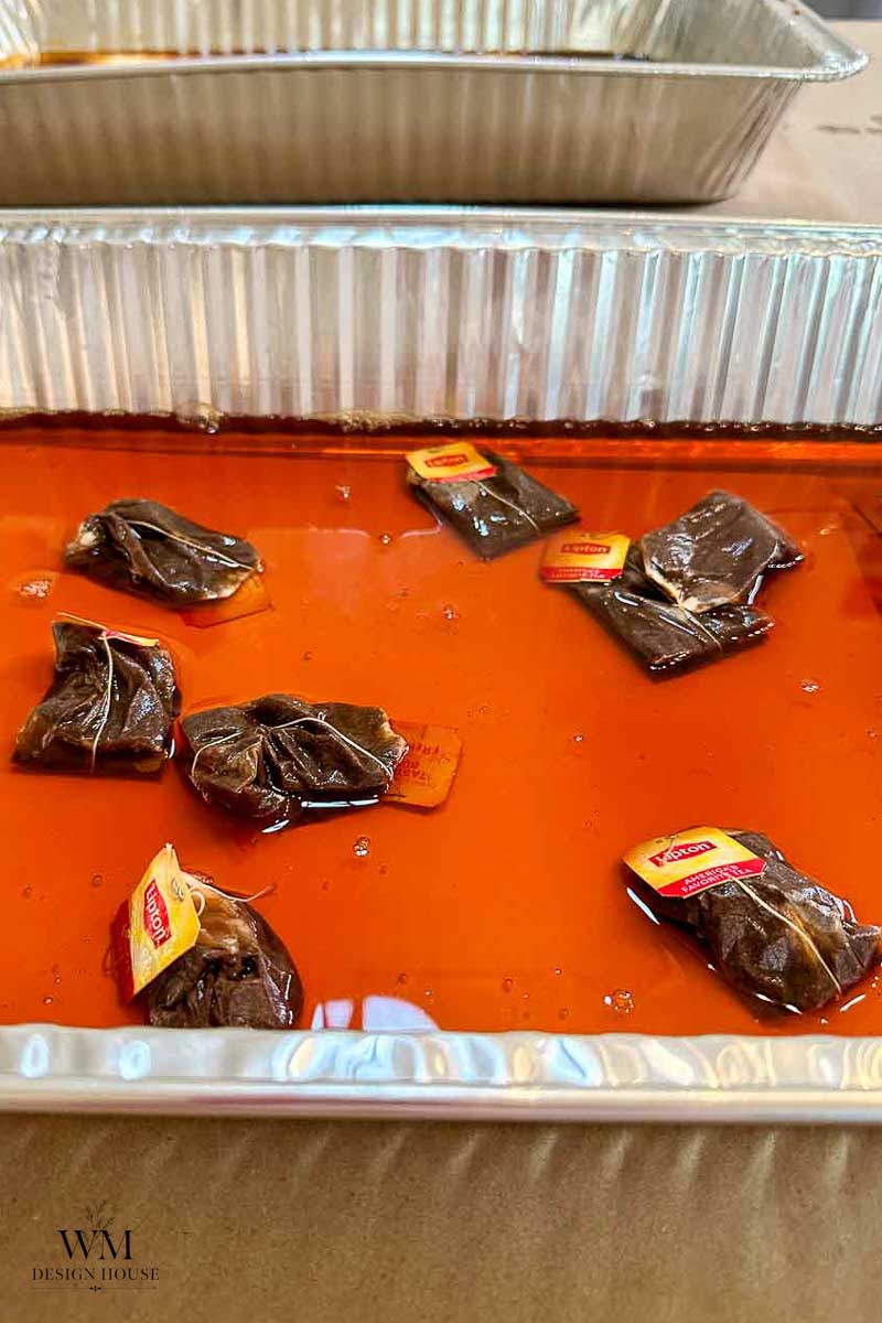 Several tea bags are soaking in orange-brown liquid inside a rectangular aluminum tray, illustrating a step often found in tutorials on how to make coffee paper. Another empty aluminum tray is visible in the background. The logo “WM Design House” appears in the bottom left corner.