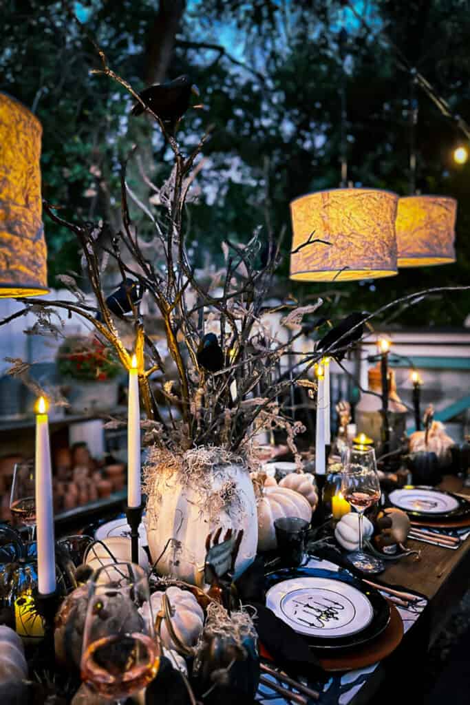 Halloween outdoor tablescape with a spooky tree and crows as the centerpiece. Shades of gray illuminate the table with lots of pumpkins and rusty elegance. Dishes are black and white, and the name cards are held with a skeleton hand. 